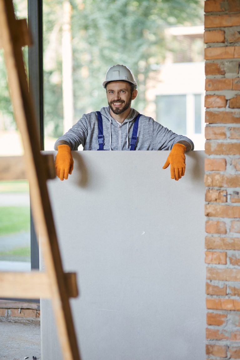 Young male builder in hard hat smiling at camera, holding drywall